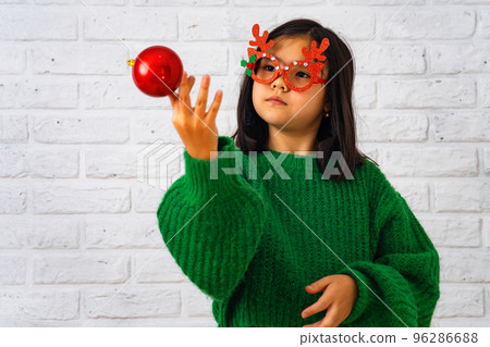 Asian girl in masquerade glasses with reindeer horns, playing with a Christmas ball Asian girl in masquerade glasses with reindeer horns, playing with a Christmas ball 96286688