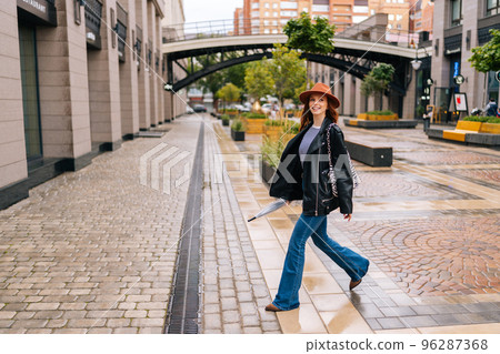 Full length of positive redhead woman wearing fashion hat walking on European city street holding in hand closed transparent umbrella. Concept of modern female lifestyle at autumn season. Full length of positive redhead woman wearing fashion hat walking on European city street holding in hand closed transparent umbrella. Concept of modern female lifestyle at autumn season. 96287368