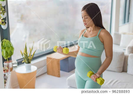 Pregnancy fitness. Pregnant woman home workout fitness strength training exercise with dumbbell weights. Body workout arm curls showing of belly and arms. 2nd and 3rd trimester workouts Pregnancy fitness. Pregnant woman home workout fitness strength training exercise with dumbbell weights. Body workout arm curls showing of belly and arms. 2nd and 3rd trimester workouts 96288893