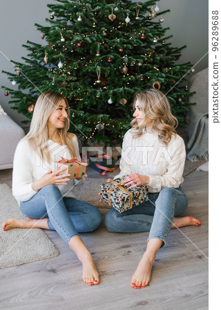 Sisterhood friends in cozy clothes smile broadly and exchange Christmas gifts in a festive box under the Christmas tree. Sisterhood friends in cozy clothes smile broadly and exchange Christmas gifts in a festive box under the Christmas tree. 96289688