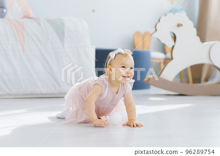 A pretty blue eyed blonde one year old in a pink dress crawls on all fours in the nursery on the floor and smiles fervently A pretty blue eyed blonde one year old in a pink dress crawls on all fours in the nursery on the floor and smiles fervently 96289754