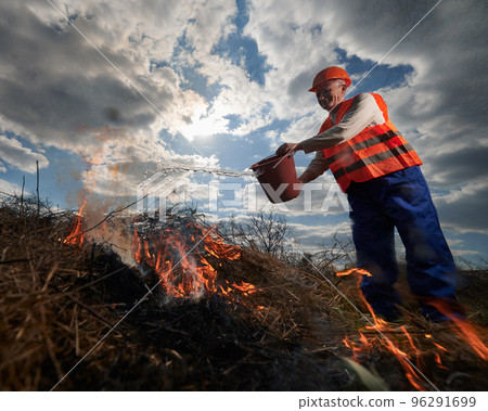 Firefighter ecologist fighting fire in field with cloudy sky on background. Male environmentalist holding bucket and pouring water on burning dry grass. Natural disaster and ecology demage concept. 96291699