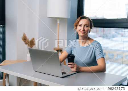 Portrait of cheerful pretty blonde woman working on laptop computer, holding cup with coffee, looking at camera, sitting at table on background of large window in light modern home office room. Portrait of cheerful pretty blonde woman working on laptop computer, holding cup with coffee, looking at camera, sitting at table on background of large window in light modern home office room. 96292175