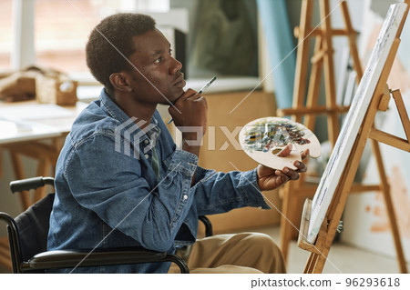 Young pensive African American painter with paintbrush sitting in wheelchair 96293618