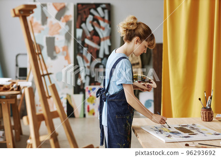 Side view of young blond woman in denim apron painting artwork 96293692