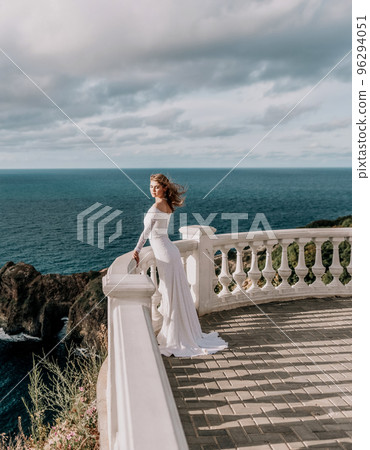 Romantic bride, blond girl in white wedding dress with open shoulders posing on open terrace with backdrop of the sea and rocks. Stylish young woman standing on terrace and looking on ocean at sunset Romantic bride, blond girl in white wedding dress with open shoulders posing on open terrace with backdrop of the sea and rocks. Stylish young woman standing on terrace and looking on ocean at sunset 96294051