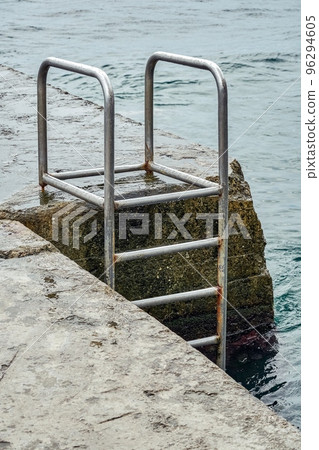 Grey concrete pier with metal ladder at blue sea water with small waves on an overcast summer day 96294605