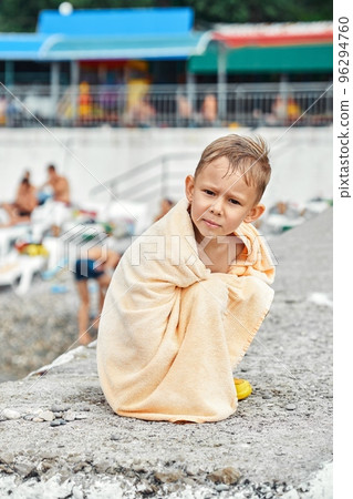 Preschooler boy covered with beige towel sit on pier and gets cold after swimming in sea. Child tries to warm-up with big terry towel sitting on concrete pier Preschooler boy covered with beige towel sit on pier and gets cold after swimming in sea. Child tries to warm-up with big terry towel sitting on concrete pier 96294760
