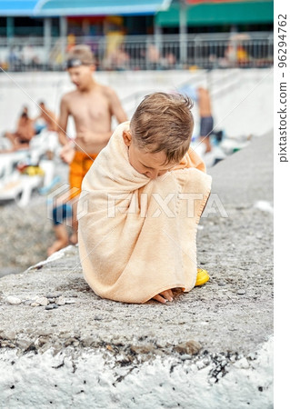 Preschooler boy covered with beige towel sit on pier and gets cold after swimming in sea. Child tries to warm-up with big terry towel sitting on concrete pier 96294762