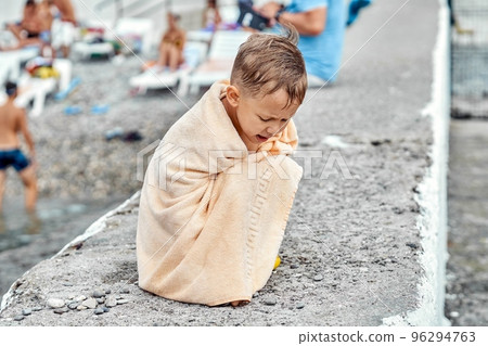 Preschooler boy covered with beige towel sit on pier and gets cold after swimming in sea. Child tries to warm-up with big terry towel sitting on concrete pier 96294763