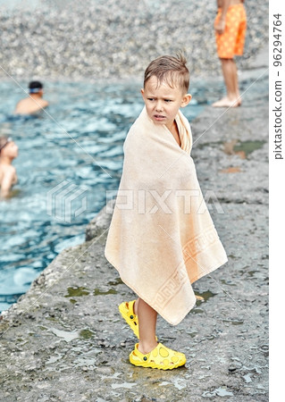Small boy wrapped in beige terry towel shivers from cold after swimming in sea. Preschooler boy tries to warm up with huge towel on stone pier closeup 96294764
