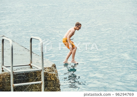 Schoolboy jumps from stone pier to sea. Moment of boy jumping and touching water. Guy looks like magician 96294769