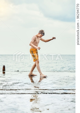 Teenager wearing yellow trunks stands on sea pier on dull day. Teen schoolboy tries to keep his balance on a wet slippery concrete pier against seascape 96294770