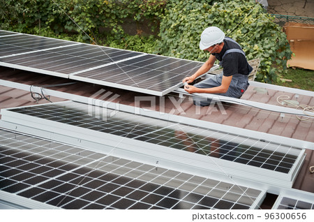Man technician mounting photovoltaic solar moduls on roof of house. Builder in helmet installing solar panel system outdoors. Concept of alternative and renewable energy. Man technician mounting photovoltaic solar moduls on roof of house. Builder in helmet installing solar panel system outdoors. Concept of alternative and renewable energy. 96300656