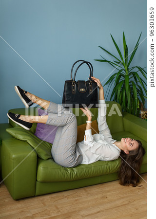 beautiful slender girl in gray plaid pants and a white blouse lying down posing on a green sofa with a small black leather handbag 96301659