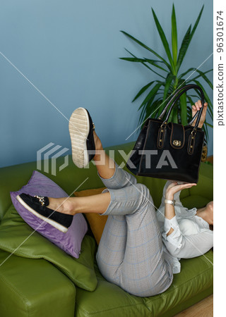 beautiful slender girl in gray plaid pants and a white blouse lying down posing on a green sofa with a small black leather handbag beautiful slender girl in gray plaid pants and a white blouse lying down posing on a green sofa with a small black leather handbag 96301674