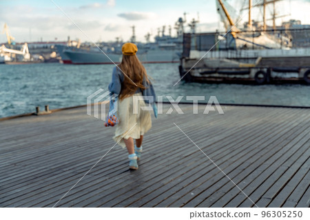 Outdoors fashion portrait of a beautiful middle aged woman walking on the beach. Marine background. Dressed in a stylish warm blue sweater, yellow skirt and beret. 96305250