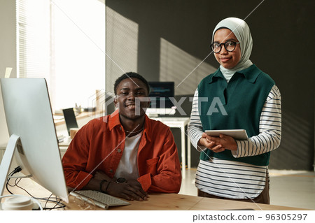 Two young software developers looking at camera while black man sitting by desk and Muslim woman with tablet standing next to him 96305297