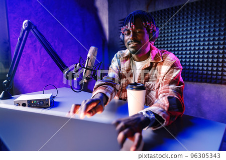 African radio host sitting at desk recording in studio with microphone and laptop African radio host sitting at desk recording in studio with microphone and laptop 96305343