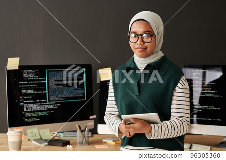 Young successful Muslim IT engineer or software developer with tablet looking at camera while standing against computer screens Young successful Muslim IT engineer or software developer with tablet looking at camera while standing against computer screens 96305360