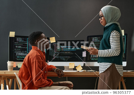 Two young confident multicultural interns in casualwear looking at each other during discussion of working points at meeting 96305377