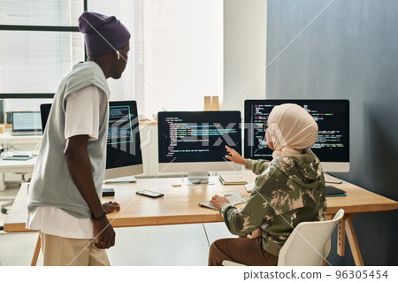 Young confident Muslim woman in hijab making presentation of decoded data on computer screen to African American male colleague 96305454