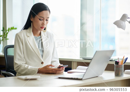 Serious elegant businesswoman working at office desk and checking text messages and notifications on smartphone 96305553