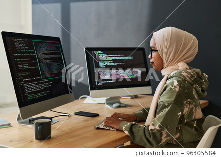 Side view of young Muslim female programmer looking through coded information on computer screen while sitting by workplace 96305584
