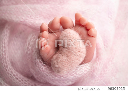 Knitted pink heart in the legs of a baby. Soft feet of a new born in a pink wool blanket. Close-up of toes, heels and feet of a newborn. Macro photography the tiny foot of a newborn baby.  96305915