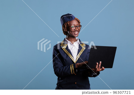 Smiling african american flight attendant wears uniform using laptop during break time at work. Stewardess checking emails from notebook looks happy for good news, studio shot. Smiling african american flight attendant wears uniform using laptop during break time at work. Stewardess checking emails from notebook looks happy for good news, studio shot. 96307721
