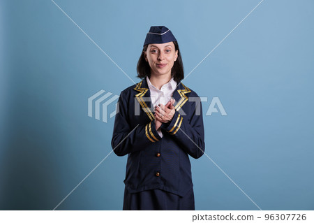 Potrait of smiling flight attendant clapping hands while standing in studio with blue background. Stewardess wearing aviation uniform applauding, shaking palms, medium shot Potrait of smiling flight attendant clapping hands while standing in studio with blue background. Stewardess wearing aviation uniform applauding, shaking palms, medium shot 96307726