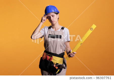 Female constructor dealing with severe headache and holding leveler on camera, rubbing temples. Woman renovation expert with painful migraine dressed with overalls and hardhat in studio. 96307761