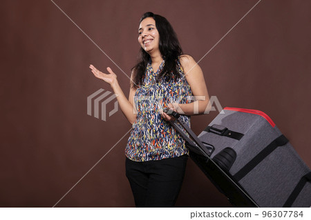 Smiling indian traveler standing with rolling suitcase, chatting, going on vacation. Woman holding baggage on wheels, cheerful tourist talking, carrying luggage in airport, trip concept Smiling indian traveler standing with rolling suitcase, chatting, going on vacation. Woman holding baggage on wheels, cheerful tourist talking, carrying luggage in airport, trip concept 96307784