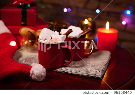 Food still life. Red cups of hot chocolate cocoa drink with marshmallows and cinnamon, on festive Christmas table against blurred background of lit candles, Xmas presents and sparkling lights garlands 96308440