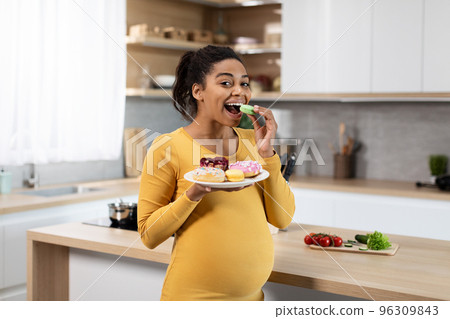 Cheerful pretty young black pregnant woman with big belly eats sweets, enjoys cookies alone Cheerful pretty young black pregnant woman with big belly eats sweets, enjoys cookies alone 96309843