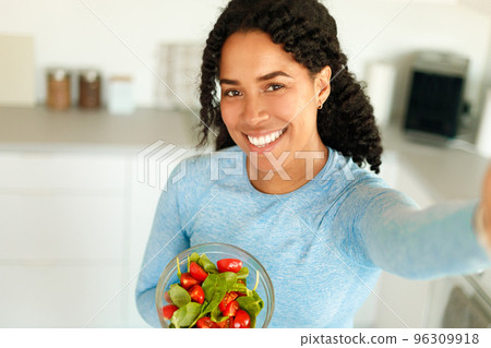 Happy african american lady holding bowl with vegetable salad and taking selfie in kitchen, recommending healthy eating Happy african american lady holding bowl with vegetable salad and taking selfie in kitchen, recommending healthy eating 96309918