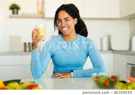 Happy black lady drinking orange juice and smiling at camera, sitting at kitchen and enjoying a well-balanced lunch Happy black lady drinking orange juice and smiling at camera, sitting at kitchen and enjoying a well-balanced lunch 96309939