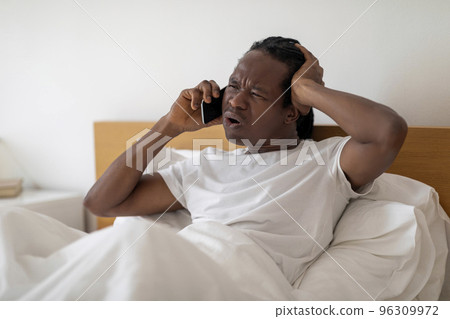 Frustrated Black Man Talking On Cellphone While Sitting In Bed At Home Frustrated Black Man Talking On Cellphone While Sitting In Bed At Home 96309972