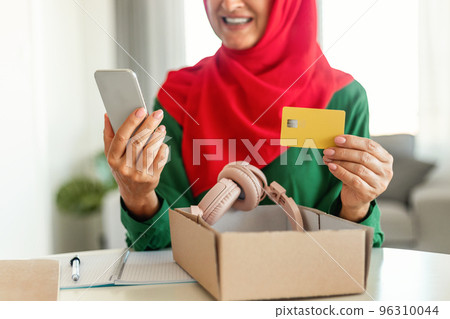 Modern mature muslim woman holding credit card and cellphone, unpacking cardboard box with new headphones 96310044