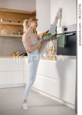 Sportive woman in grey leggins and top in a kitchen. Woman is standing on a twine and holding a plate with fresh vegetables. Healthy diet, sport concept. 96310746