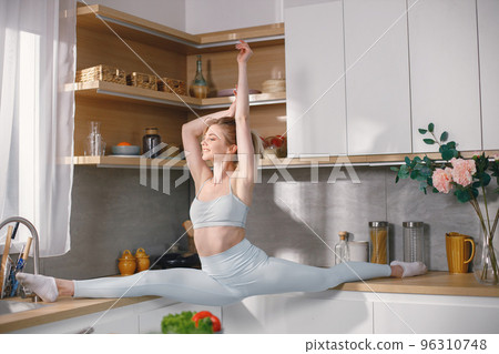 Sportive woman in grey leggins and top in a kitchen. Woman is sitting on a twine. Healthy diet, sport concept. Sportive woman in grey leggins and top in a kitchen. Woman is sitting on a twine. Healthy diet, sport concept. 96310748