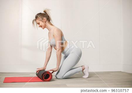 Sporty young woman rolling a red mat on a floor and posing for a photo. Woman wearing grey top and leggins. Woman after training. 96310757