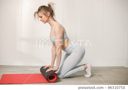 Sporty young woman rolling a red mat on a floor and posing for a photo. Woman wearing grey top and leggins. Woman after training. Sporty young woman rolling a red mat on a floor and posing for a photo. Woman wearing grey top and leggins. Woman after training. 96310758