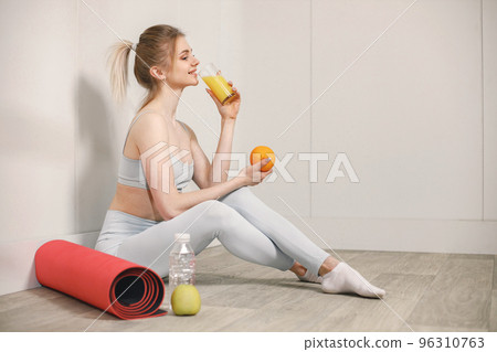 Sporty young woman sitting on a floor with glass of juice and posing for a photo. Woman wearing grey top and leggins. Woman sitting near red mat and bottle of water, an orange and an apple. 96310763