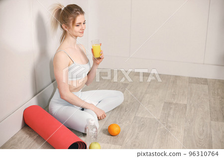 Sporty young woman sitting on a floor with glass of juice and posing for a photo. Woman wearing grey top and leggins. Woman sitting near red mat and bottle of water, an orange and an apple. Sporty young woman sitting on a floor with glass of juice and posing for a photo. Woman wearing grey top and leggins. Woman sitting near red mat and bottle of water, an orange and an apple. 96310764
