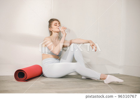 Sporty young woman sitting on a floor and posing for a photo. Woman wearing grey top and leggins. Woman sitting near red mat and drink a water. 96310767