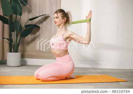 Sporty young woman training with elastic rope at home. Woman wearing rose top and leggins. Woman doing exercises on an orange mat. 96310784