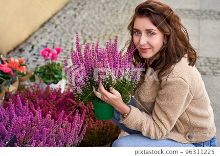 Young beautiful woman holding a pot with blooming violet heather plant in a street flower shop. Young beautiful woman holding a pot with blooming violet heather plant in a street flower shop. 96311225