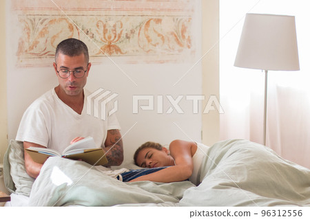Focused man reading book in morning while his wife sleeping 96312556