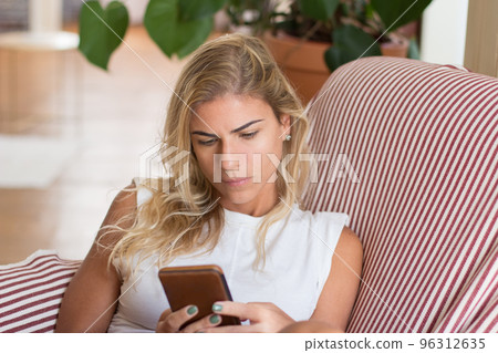 Pensive woman sitting in armchair and looking at phone screen 96312635